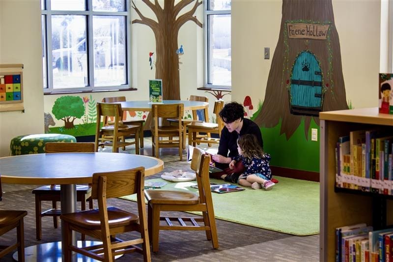 An adult and a child sit on a green rug reading books together in a colorful, forest-themed children's library area with wooden chairs, tables, and bookshelves.