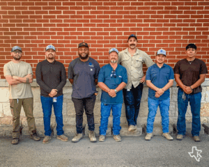 Seven men wearing work clothes and boots stand in a row, smiling in front of a brick wall. The ground is paved, and a stone base runs along the bottom of the wall.