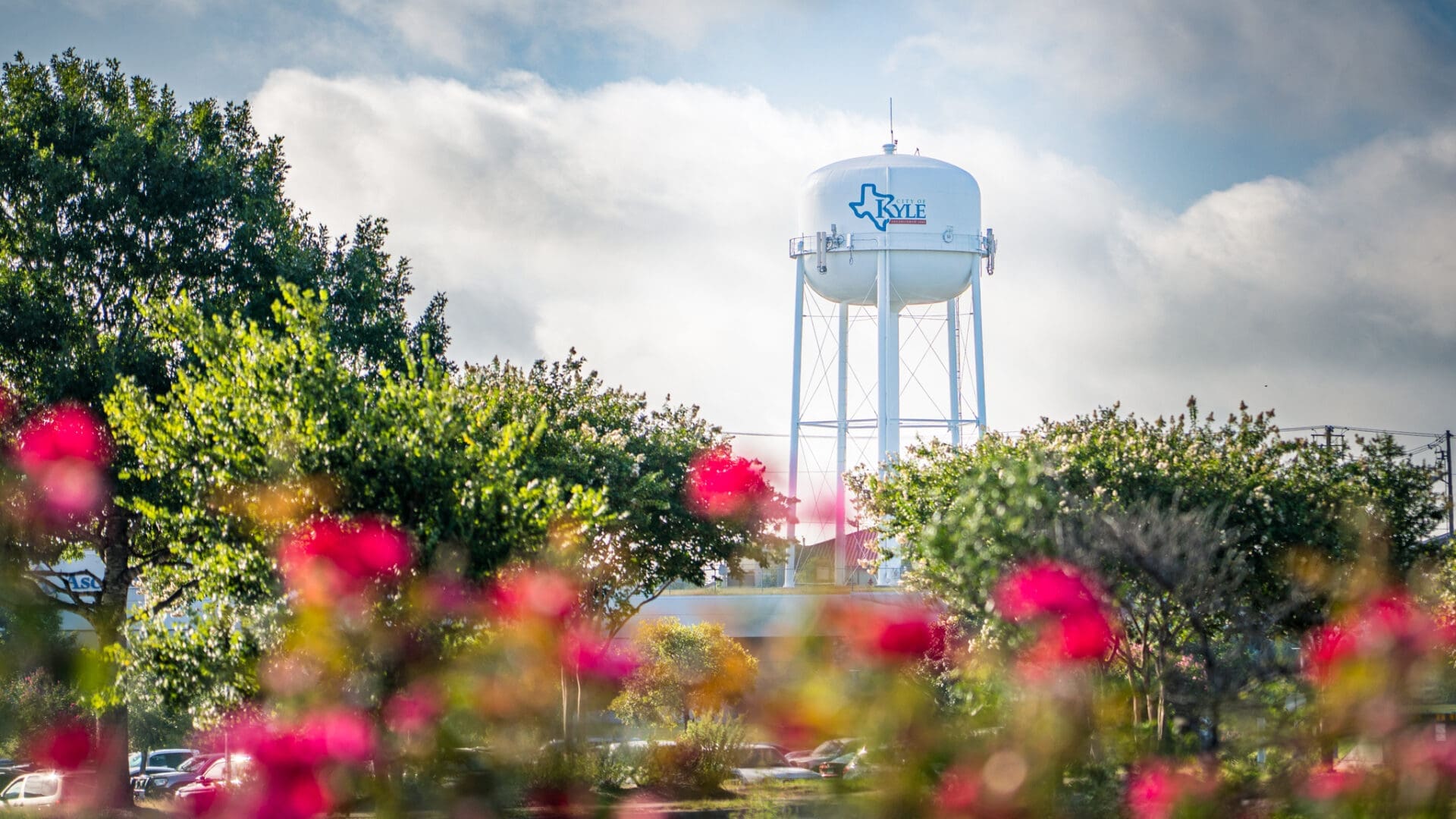 A white water tower labeled "Kyle" with a Texas outline stands tall behind blooming pink flowers and green trees under a partly cloudy sky. The water tower pictured is a planned site for new wireless telecommunication towers.