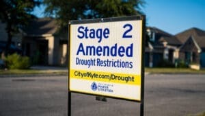 A sign in a suburban neighborhood reads, "Stage 2 Amended Drought Restrictions," with the City of Kyle Water Utilities website link below. Houses and trees are visible in the blurred background.