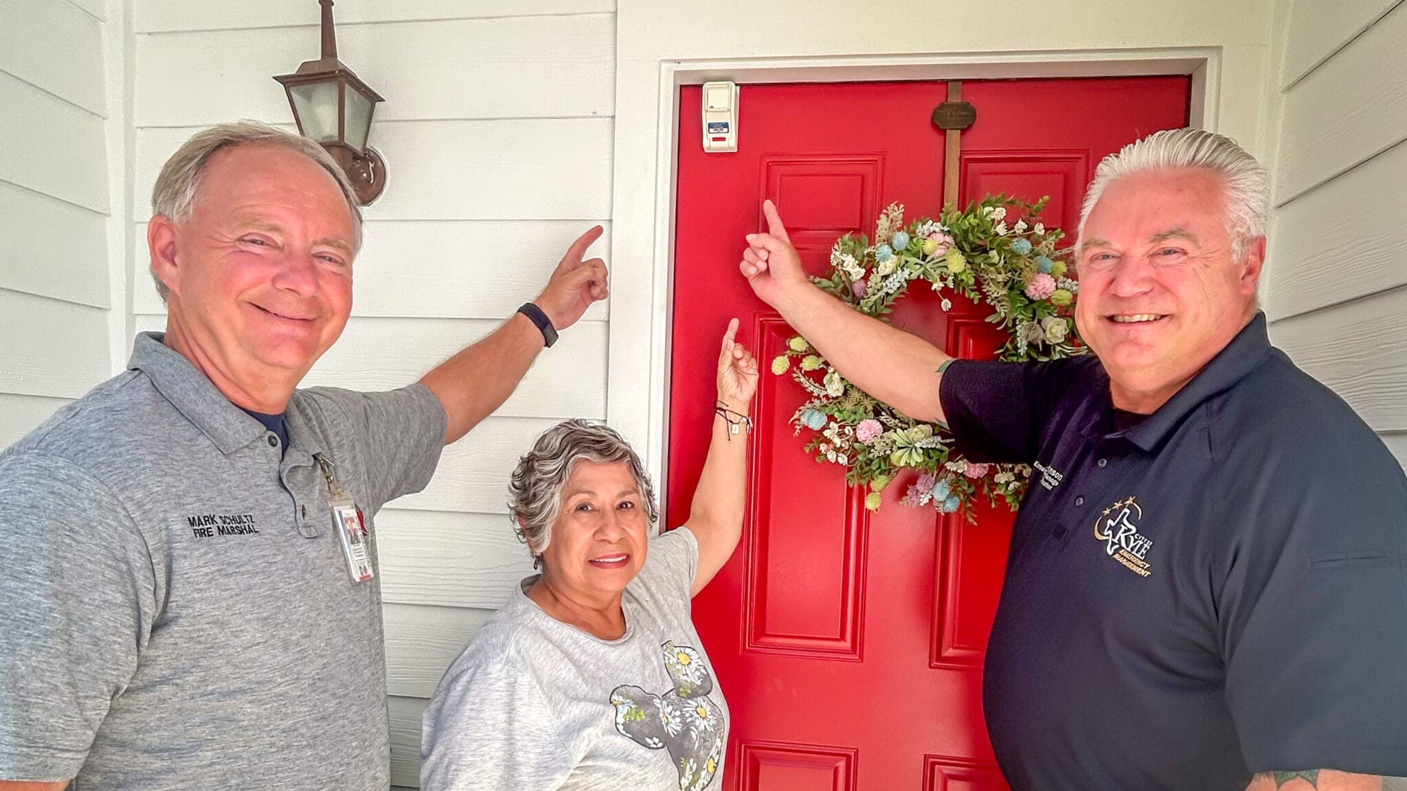 Three smiling adults stand in front of a red door with a floral wreath, pointing at a small security device, a knox box, mounted above the door. The group appears friendly and happy.