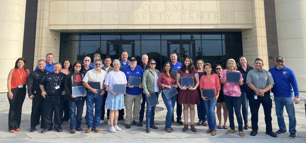 A group of people, some holding plaques or certificates, pose for a photo in front of the City of Kyle Public Safety Center building on a sunny day.