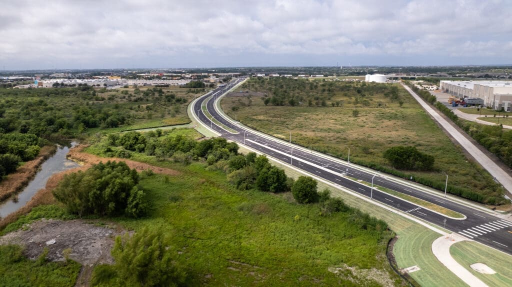 Aerial view of a newly constructed, empty road curving through green fields and sparse trees, with commercial buildings and cloudy sky in the background.
