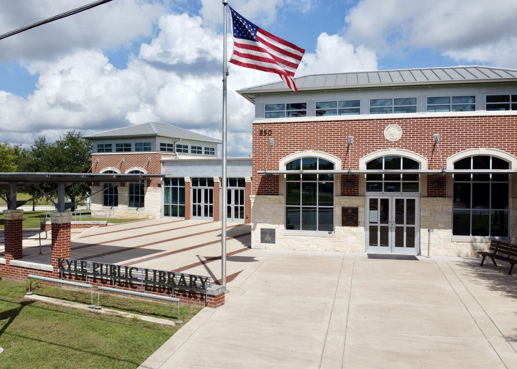 A modern brick and stone public library building with large arched windows, an American flag flying out front, and a sign reading "Public Library" near the entrance, under a partly cloudy sky.