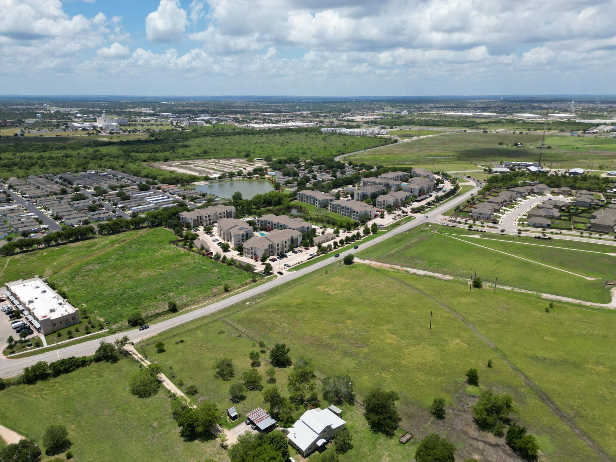Aerial view of suburban area with apartment complexes, houses, green fields, a pond, and roads under a partly cloudy sky.