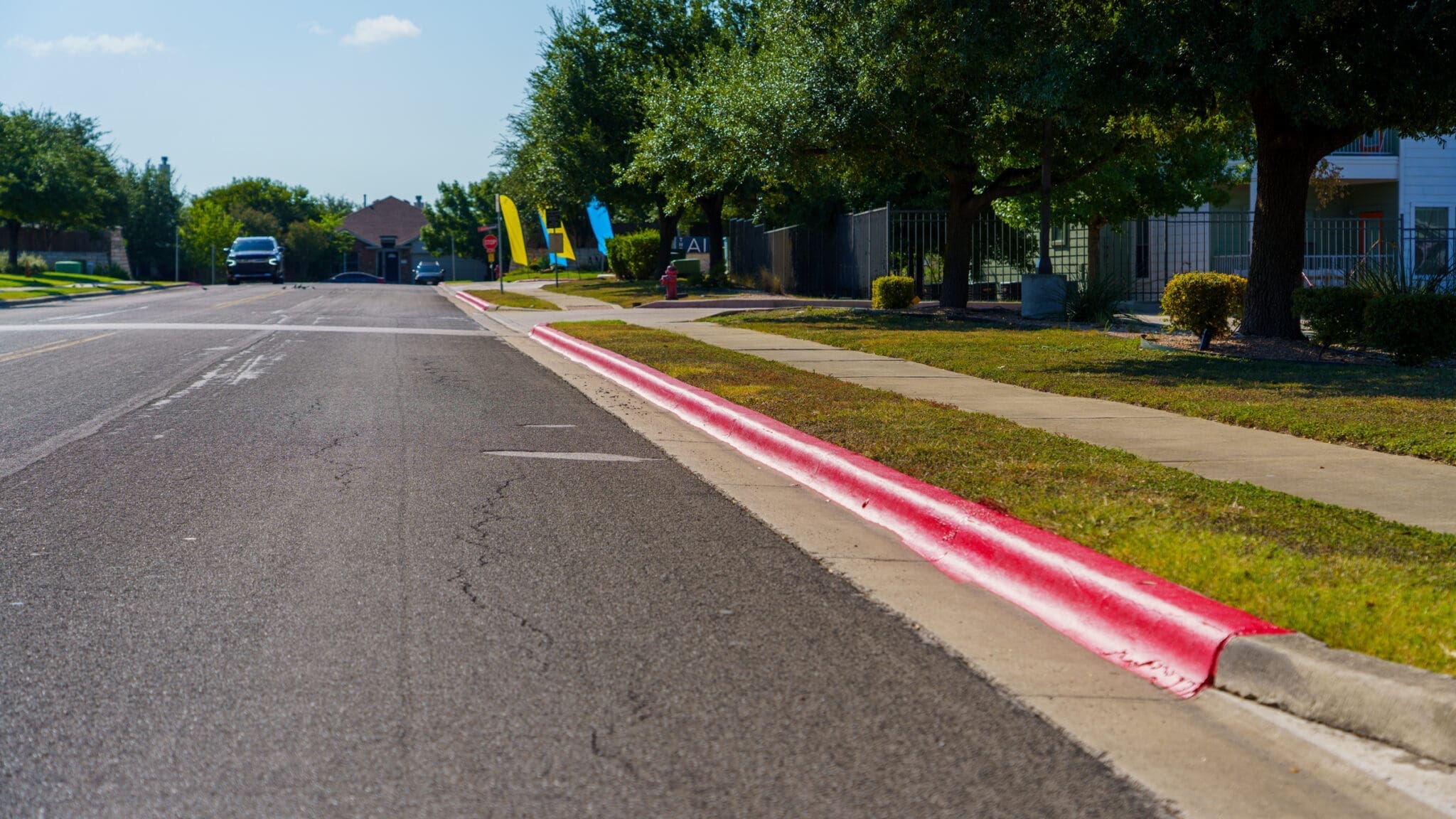 A suburban street with a sidewalk and red-painted curb, indicating no parking. Trees and houses line the right side, while colorful flags are visible in the distance under a sunny blue sky.