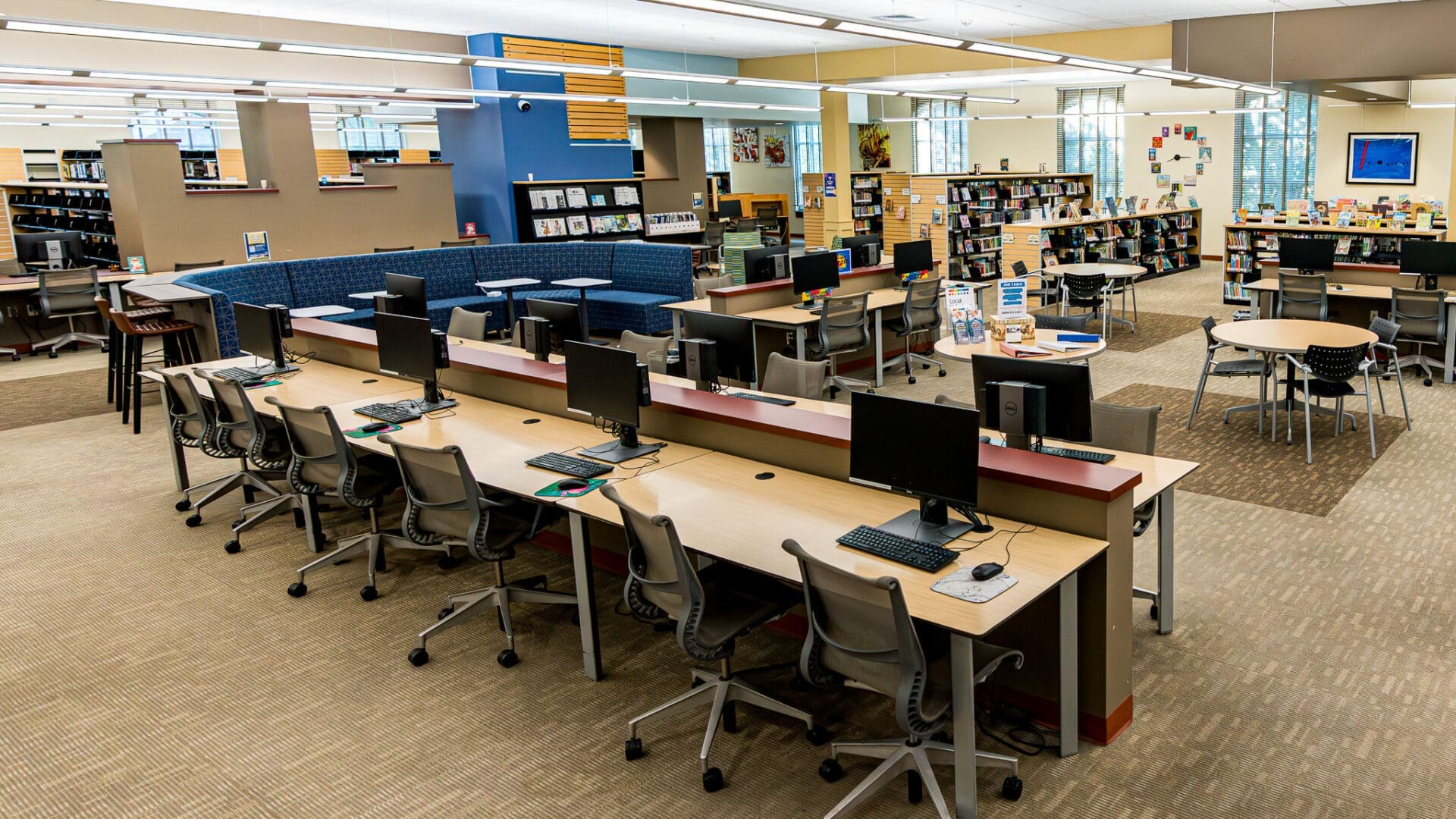 A modern library interior with rows of computer workstations, chairs, bookshelves filled with books, tables for group work, and large windows letting in natural light. The space is open, bright, and organized.