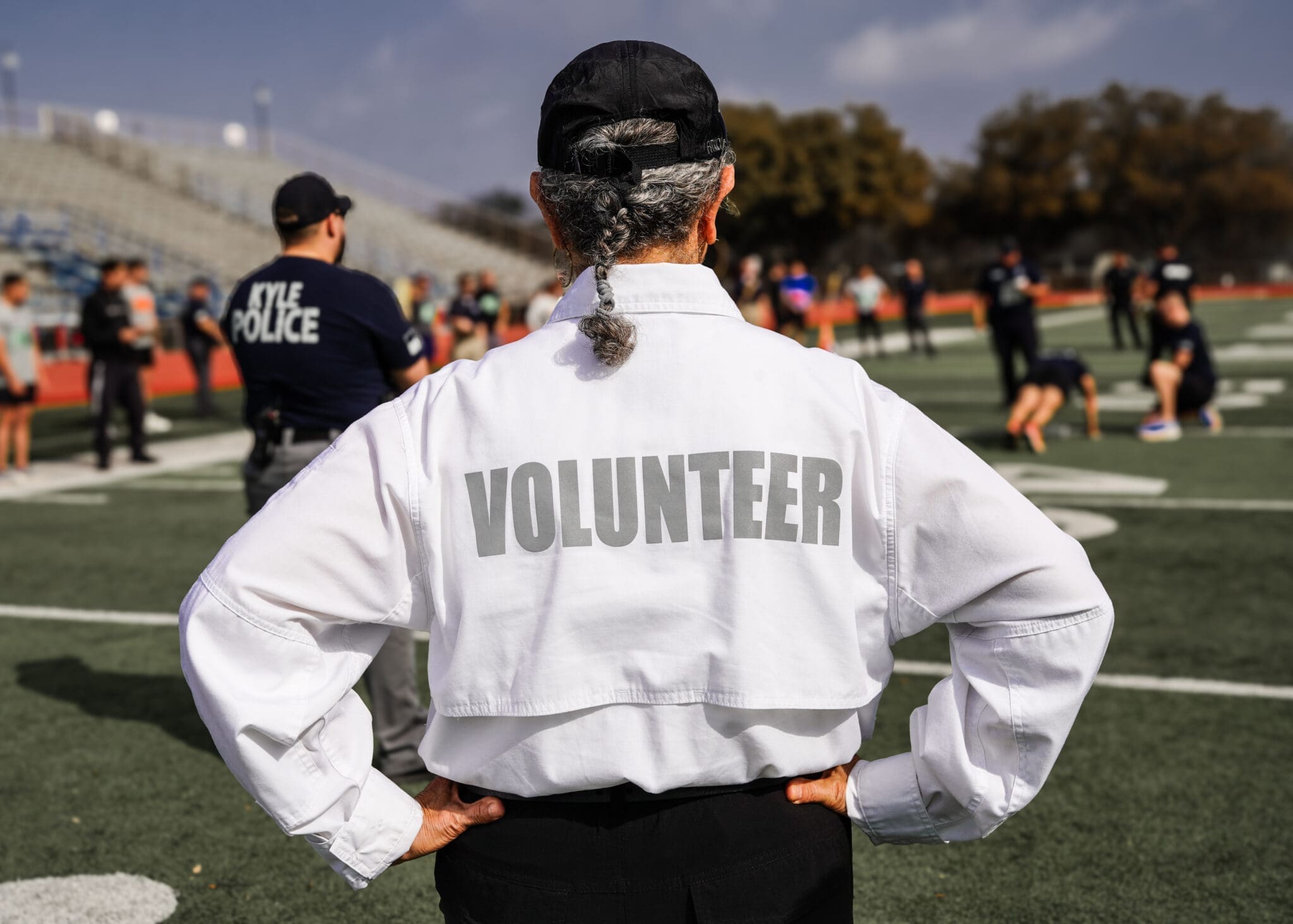 A a Citizens on Patrol member with gray hair in a braid, wearing a white shirt labeled "VOLUNTEER," stands on a sports field with hands on hips while groups of people and police officers are visible in the blurred background.