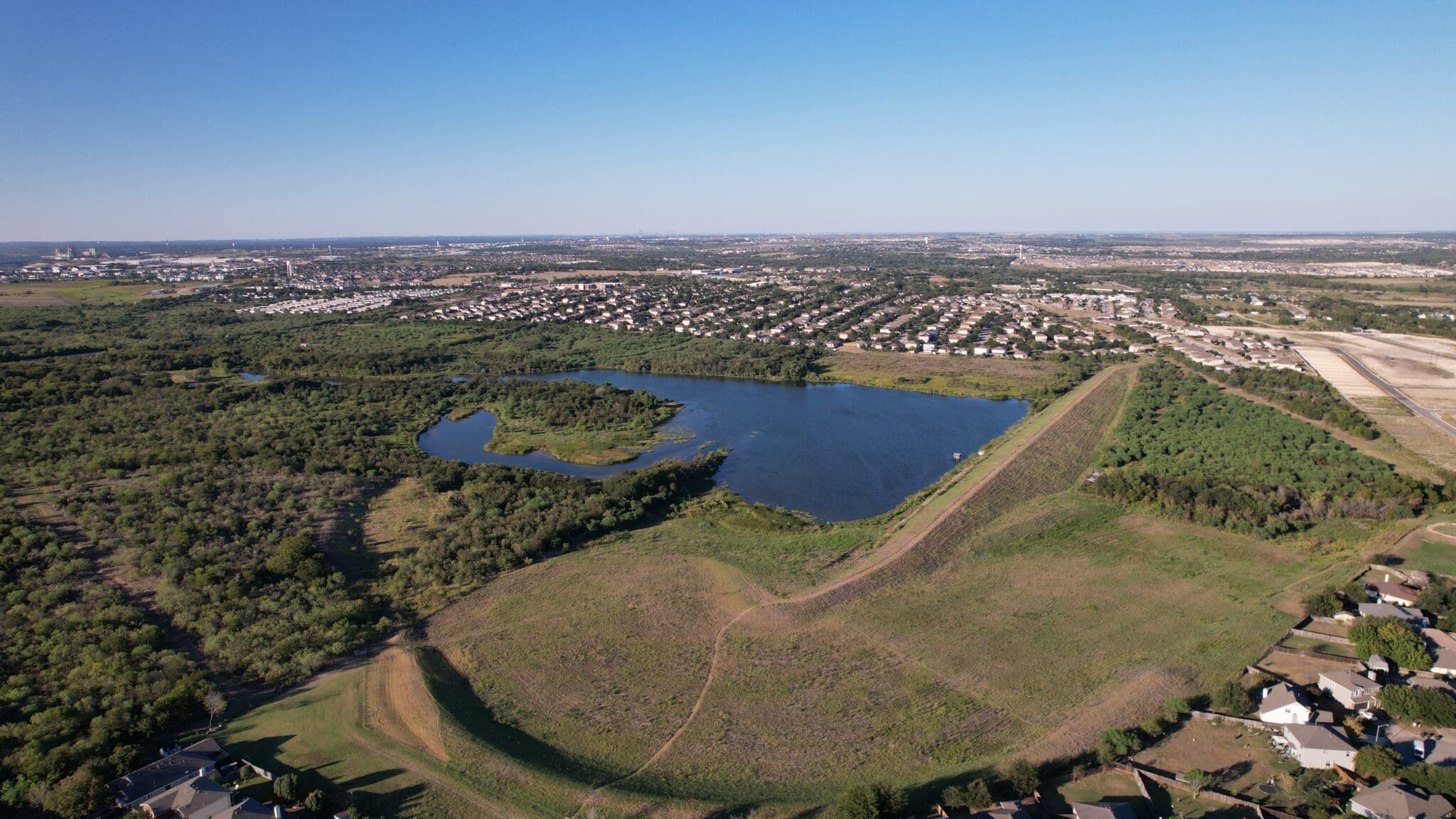 Aerial view of Lake Kensington surrounded by greenery, with clusters of trees and grassy fields. Residential neighborhoods with rows of houses border the water and extend into the distance under a clear blue sky.