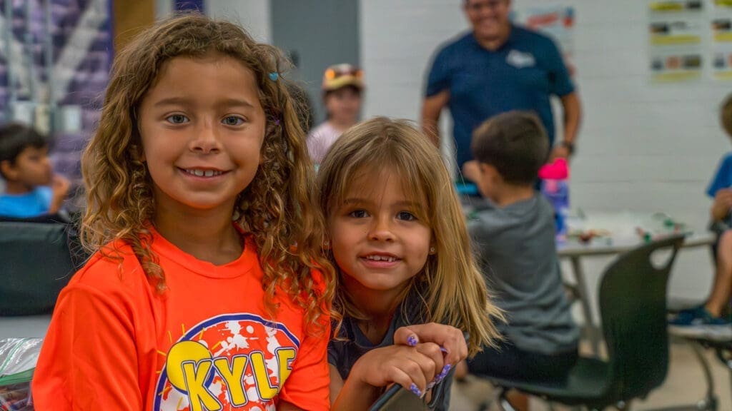 Two girls attending Kyle Summer Camp, one in a bright orange camp shirt, smile for the camera.