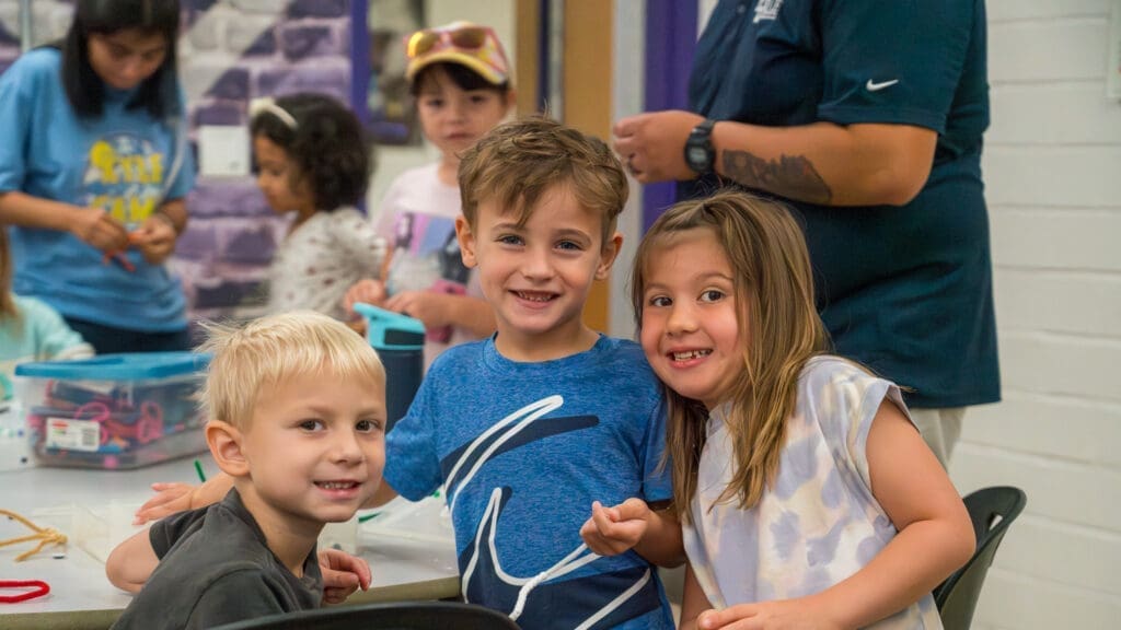 Three children smile for the camera during a Kyle Summer Camp arts and crafts activity.