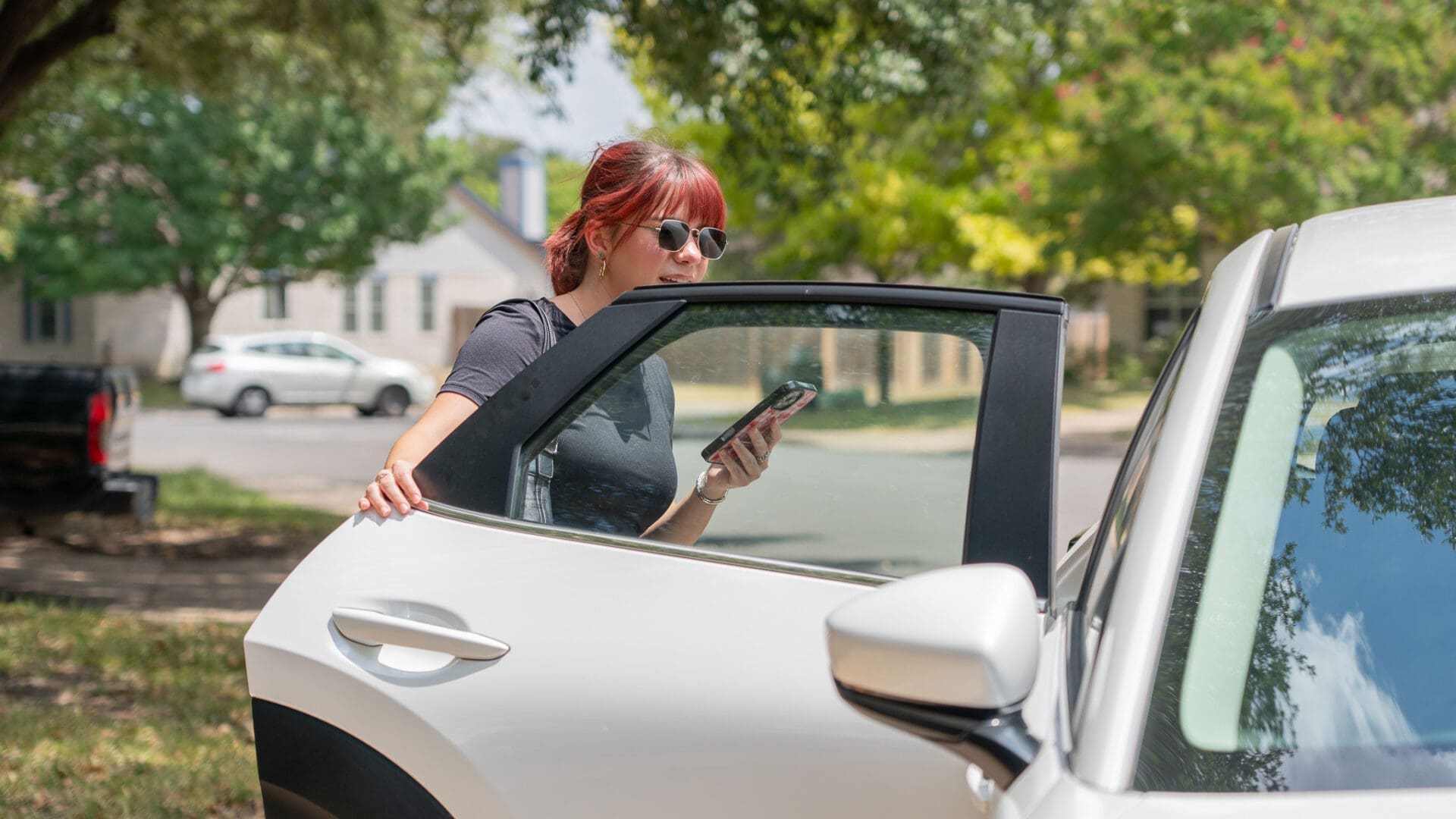A woman with red hair and sunglasses stands next to an open car door, holding her phone and looking at the screen on a sunny day, with trees and houses in the background.