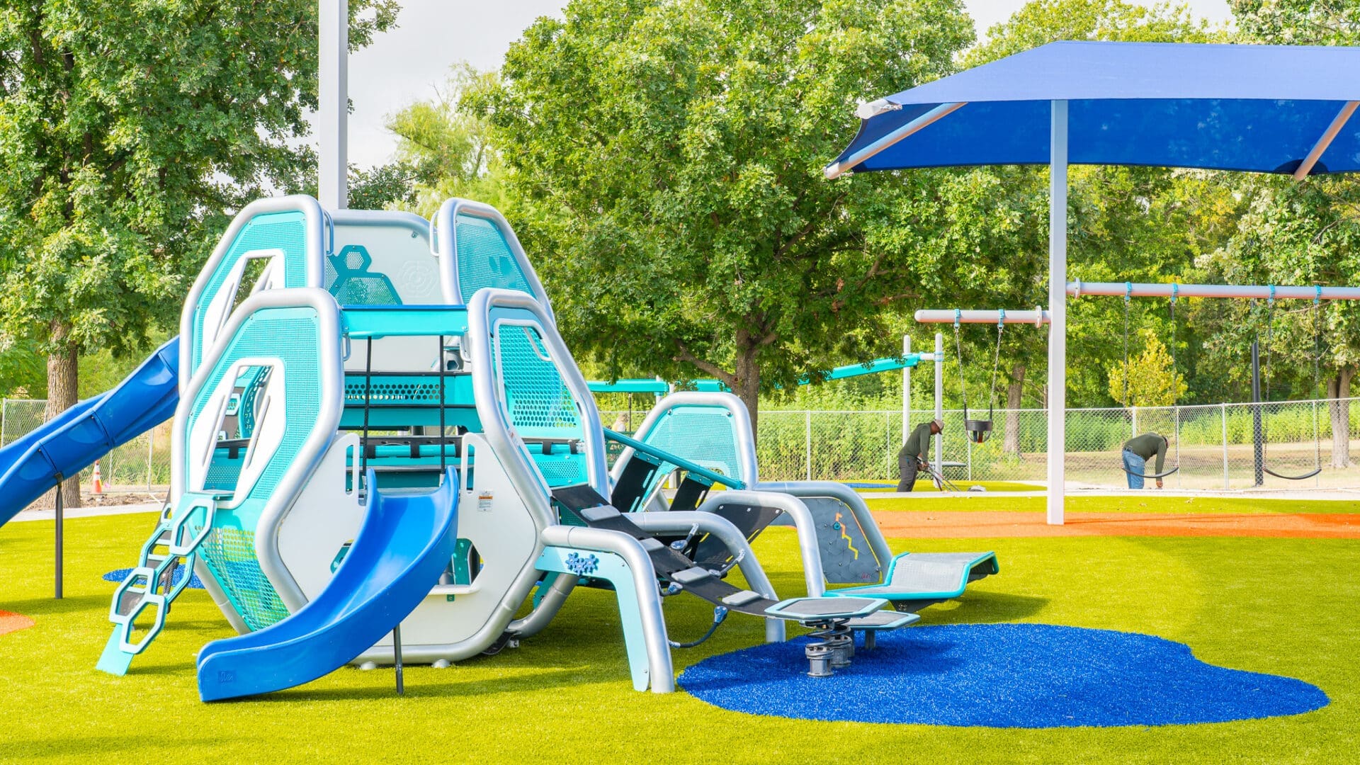 A modern playground with blue and white climbing structures, slides, and a seesaw on green artificial turf. A large blue shade canopy covers part of the area, with trees and fencing in the background.
