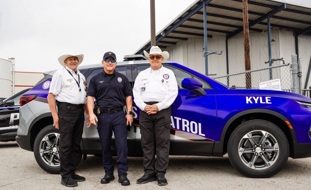 Two members of Kyle Citizens on Patrol stand with Kyle Chief of Police Jeff Barnett in front of a grey, blue and red accented SVU outdoors with a cloud sky. 
