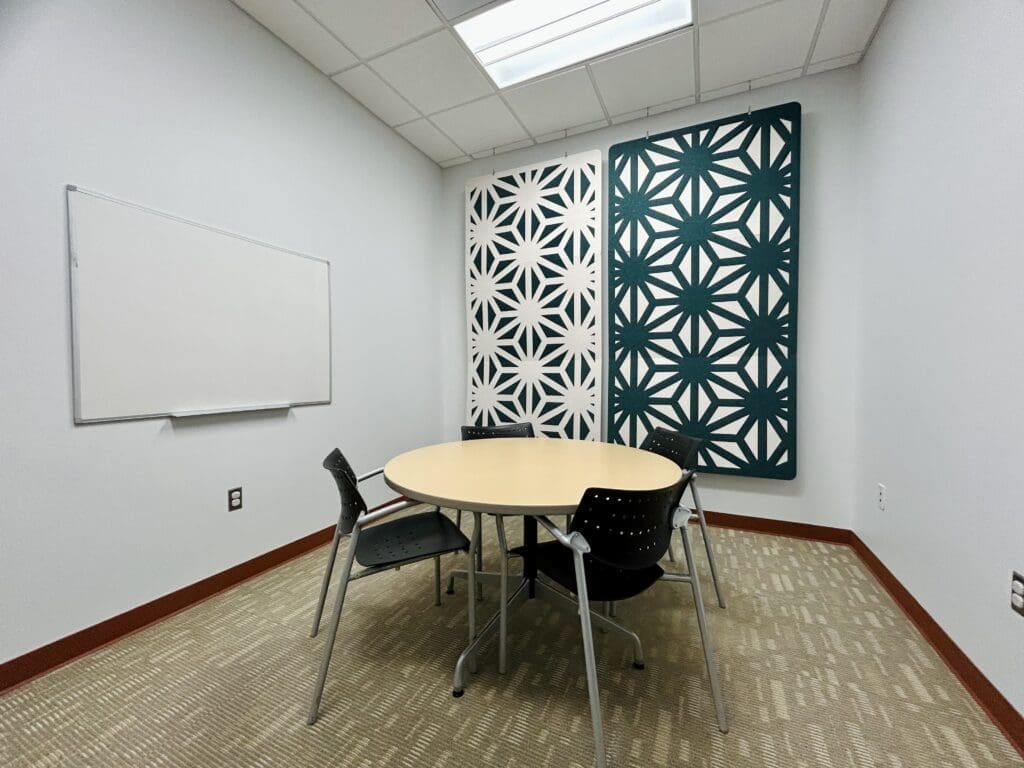 A small meeting room with a round table, four black chairs, a whiteboard on the wall, and two large panels with geometric patterns. The room has beige carpet and white walls.
