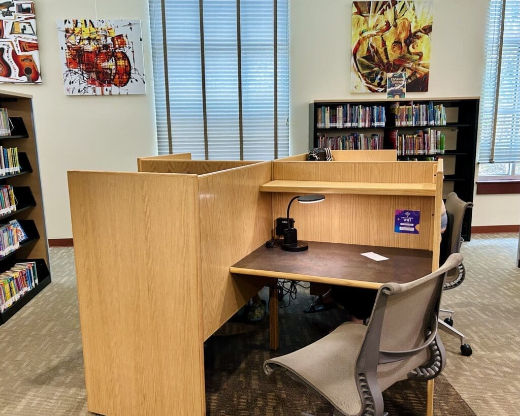A library study cubicle with a chair, lamp, and microscope on the desk. Bookshelves and colorful artwork are visible in the background, and sunlight streams through tall windows with blinds.
