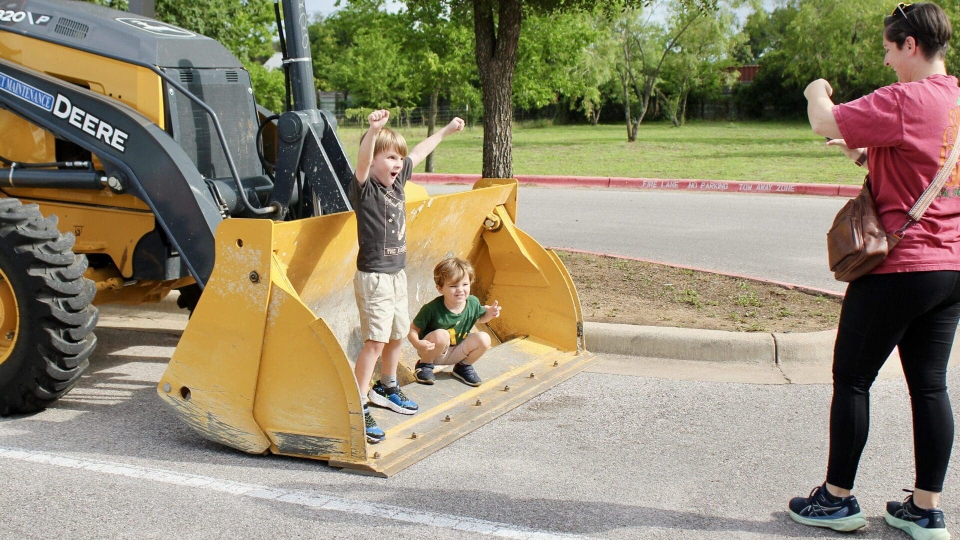 Two young children pose excitedly in the bucket of a yellow construction vehicle while a woman stands nearby taking their photo in a sunny outdoor setting with trees in the background.