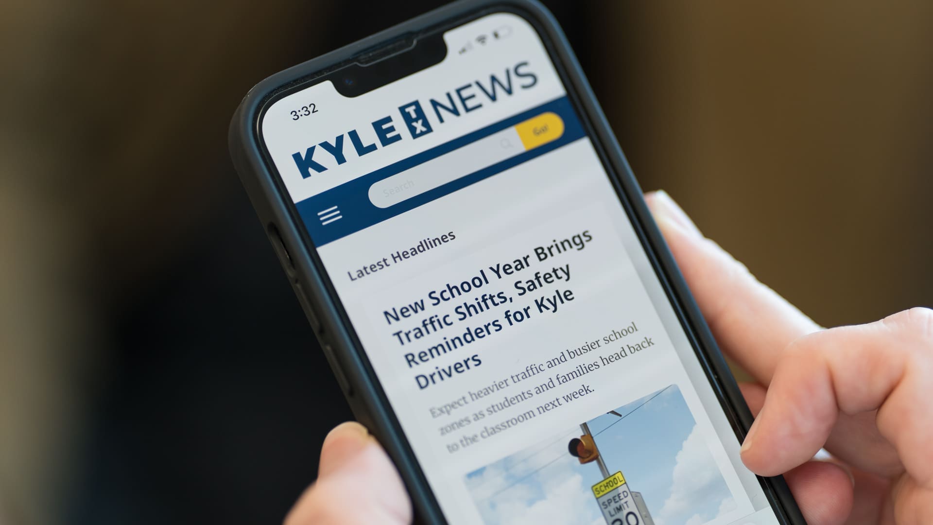 A person holds a smartphone displaying a news website called "KYLE TX NEWS," with a headline about new school year traffic shifts and safety reminders for drivers. The article is partially visible on the phone screen.