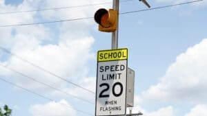 A traffic sign reads "School Speed Limit 20 When Flashing" beneath a yellow flashing light against a partly cloudy sky, indicating a reduced speed zone near a school.