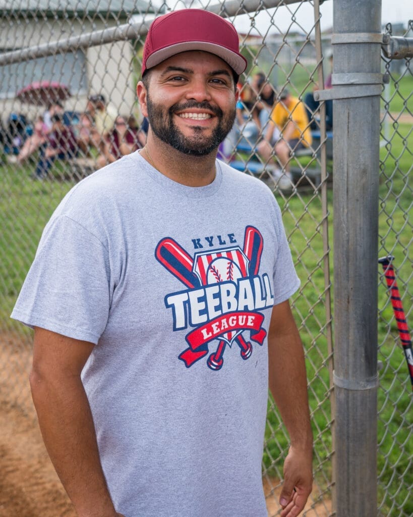 A smiling man wearing a maroon cap and a gray "Kyle Teeball League" t-shirt stands by a chain-link fence on a baseball field, with people sitting and watching in the background.