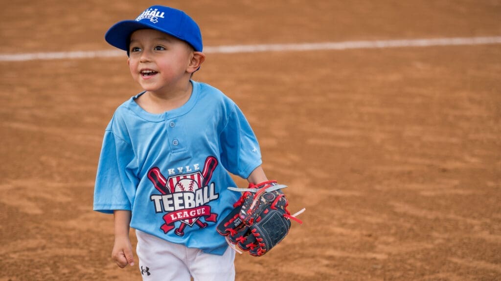 A young boy wearing a blue "TeeBall League" shirt and blue cap stands on a baseball field, smiling and holding a baseball glove.