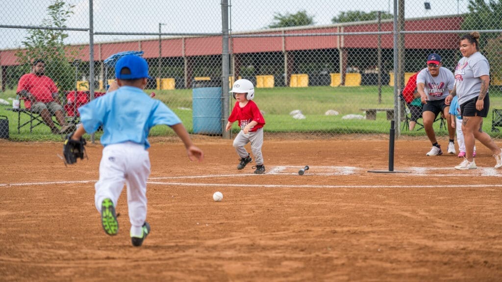 A young child in a red shirt and helmet runs toward first base after hitting a baseball off a tee, while another child in a blue uniform runs toward the ball on a baseball field. Coaches and spectators watch from the sidelines.