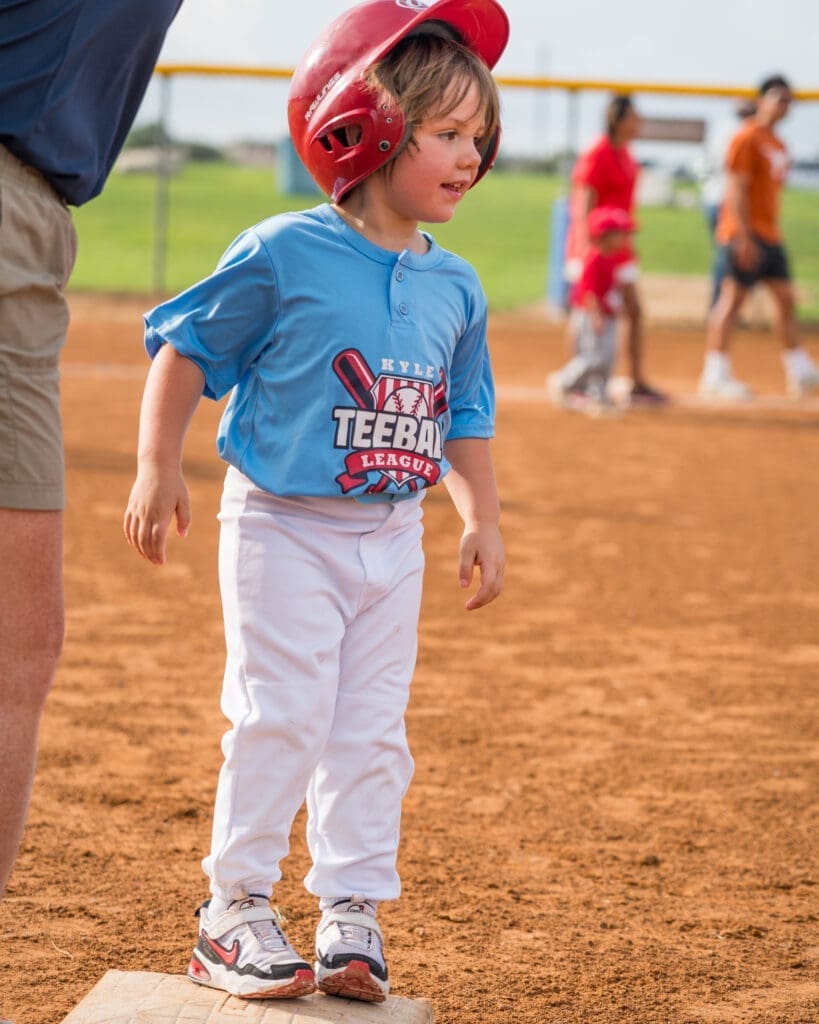 A young child wearing a red helmet and blue "Tee Ball League" shirt stands on a base during a baseball game, with other players and a coach visible in the background on a sunny day.