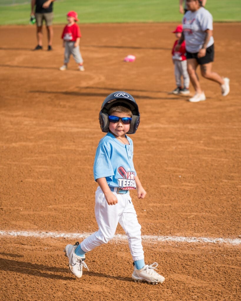 A young child in a blue baseball uniform, helmet, and sunglasses walks confidently on a baseball field. Other kids and adults are visible in the background.