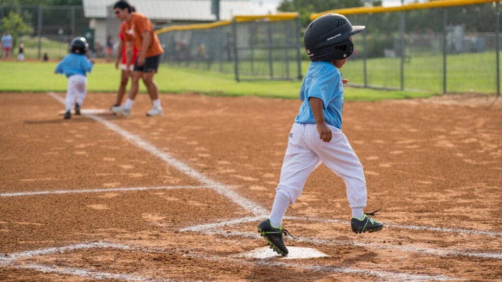 A young child in a helmet and blue shirt runs on a baseball field, approaching home plate, while another child and an adult appear in the background on a sunny day.