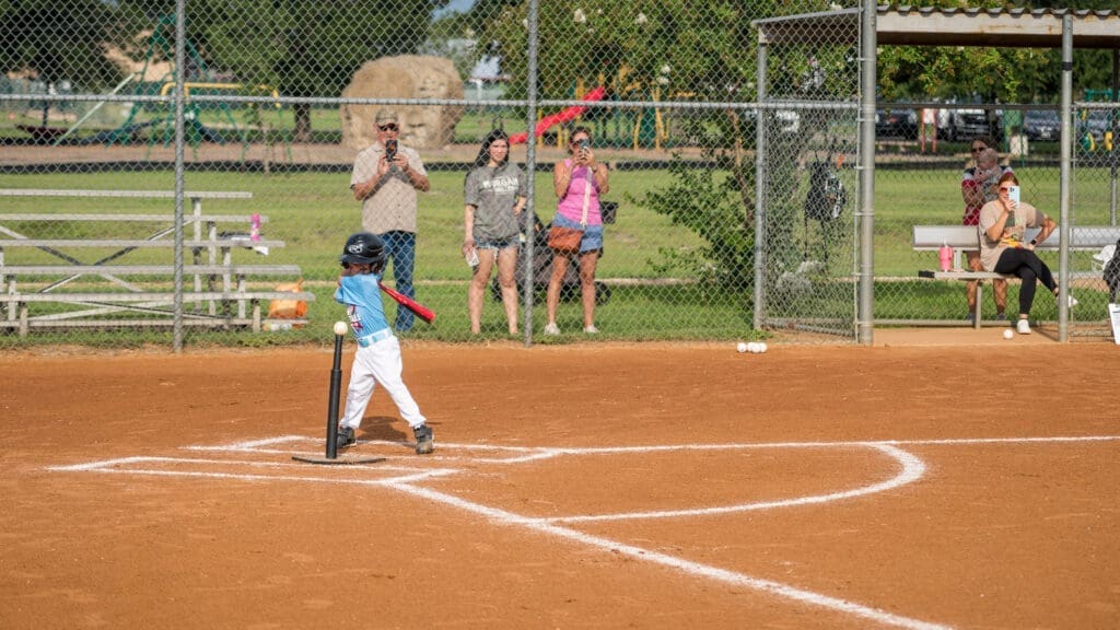 A young child in a helmet swings a bat at a baseball on a tee during a game, while adults watch and take photos from behind a chain-link fence and nearby benches on a sunny day.