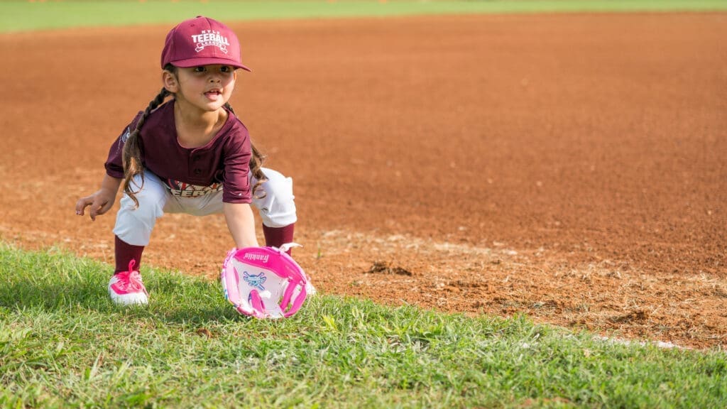 Young girl in a maroon baseball uniform and cap squats on a baseball field, reaching down with a pink glove to field a ball, with grass and dirt visible around her.