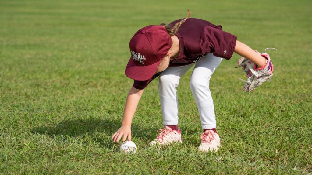 A young child in a maroon baseball uniform and cap bends down to pick up a baseball from a grassy field, holding a glove in one hand.