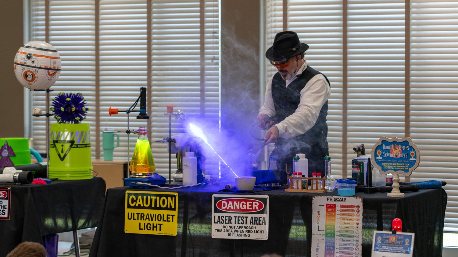 A person wearing a hat and safety goggles performs a science experiment with smoke and bright UV light at a table covered in lab equipment and caution signs as part of a demonstration.