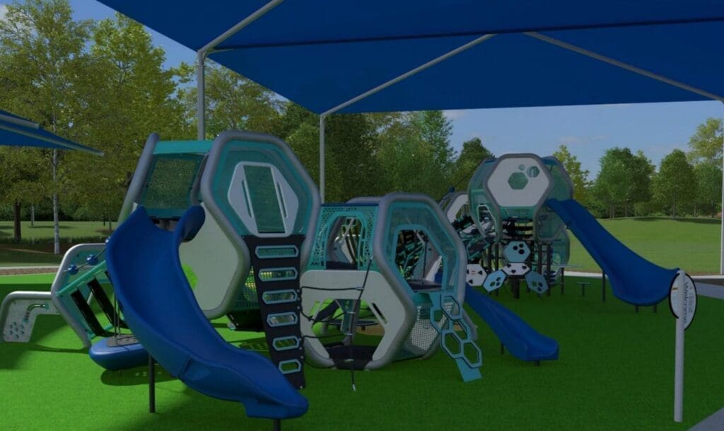 A modern playground with geometric climbing structures, blue slides, and shade canopies, set on green artificial turf with trees in the background.