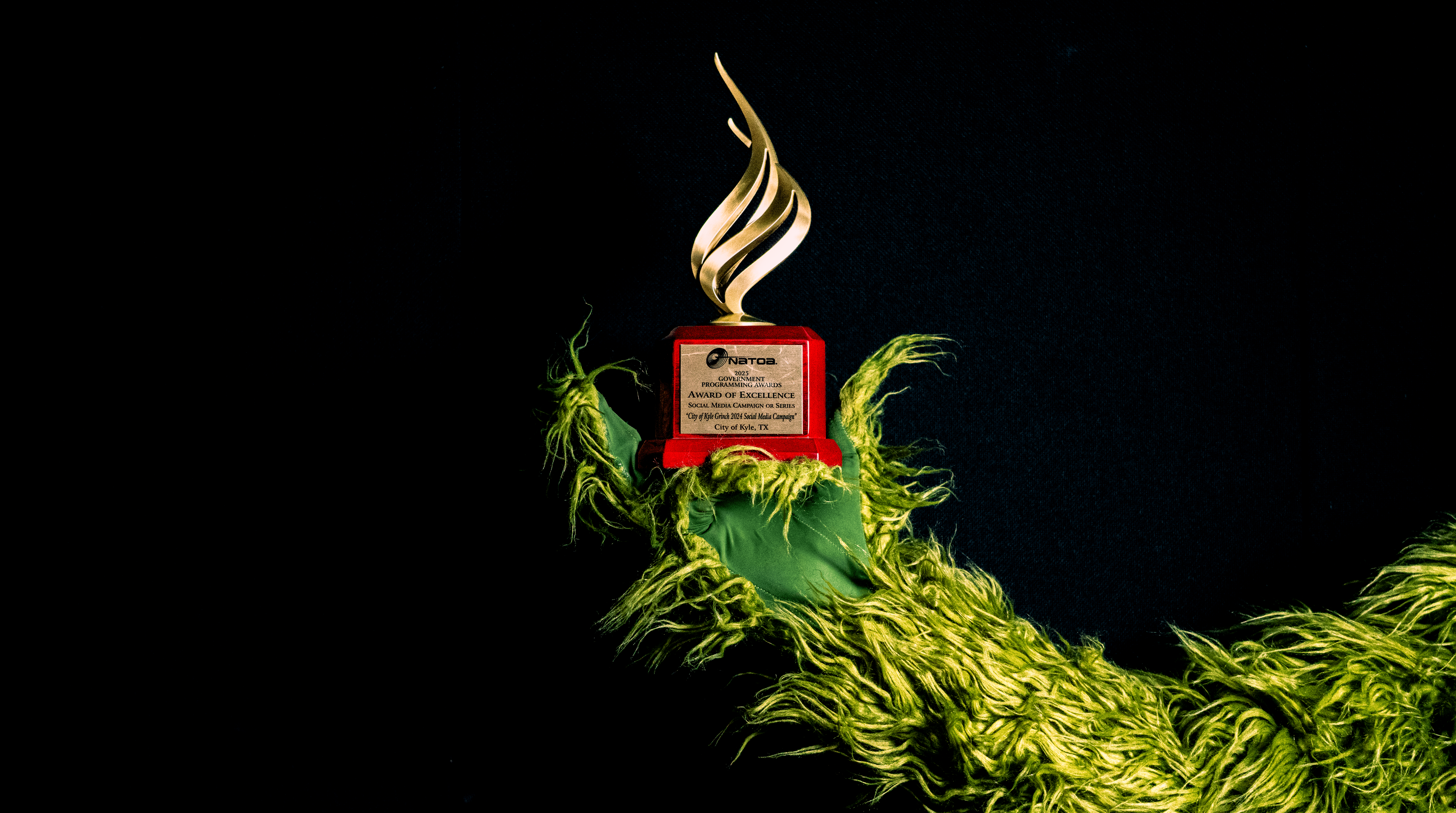A gold NATOA Award of Excellence trophy with a red base is held by a green, furry hand resembling the Grinch, against a black background.