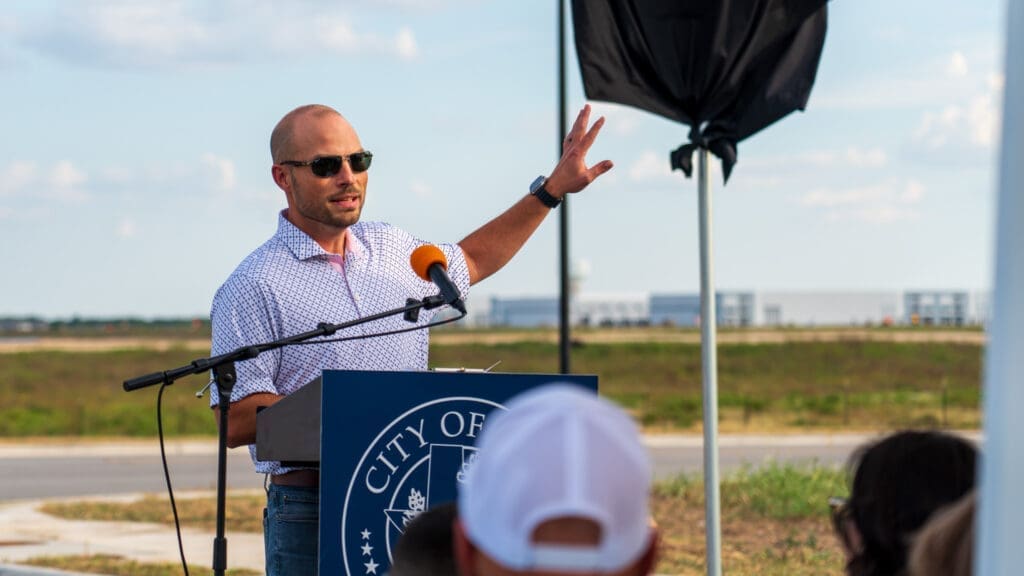 Mayor Travis Mitchell speaks at an outdoor podium with a microphone, gesturing with one hand. The podium has "City of Kyle" written on it. Several people are seated in the foreground, listening.