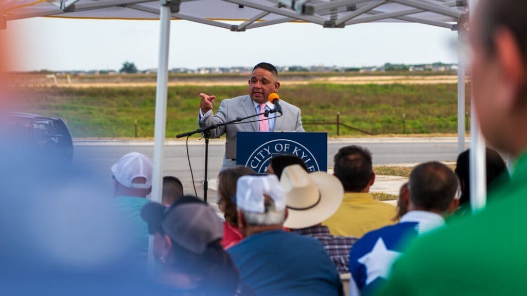 A man in a suit speaks at a podium with "City of Kyle" on it, addressing an outdoor audience seated under tents. Some audience members wear hats, and a Texas flag design is visible on one shirt.
