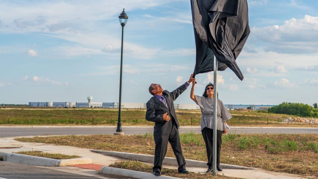 A man in a suit and a woman in sunglasses unveil a sign covered by a black cloth on a street corner, with a grassy field, sidewalk, and industrial buildings in the background.
