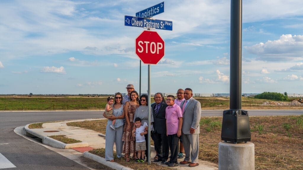 A group of people, including children, pose and smile at a street corner next to a stop sign and blue street signs reading “Logistics” and “Chevo Pastrano Sr.” A wide, open field stretches out behind them under a blue sky.