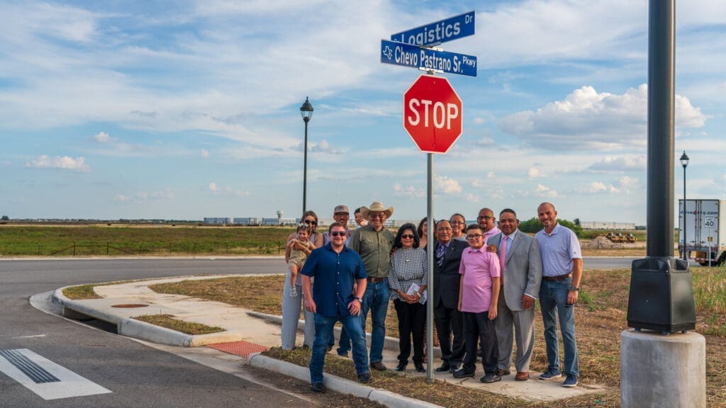 A group of people pose together at a street intersection by a stop sign and street signs for Logistics Dr and Chevo Pastrano Sr Pkwy, with an open field and blue sky in the background.