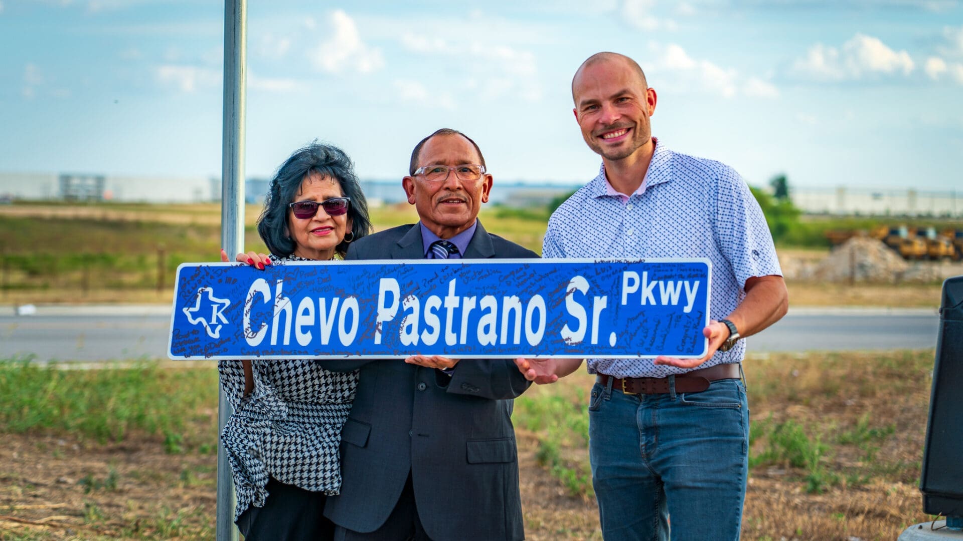 Three people stand outdoors smiling and holding a blue street sign that reads "Chevo Pastrano Sr. Pkwy" with a Texas logo. The background shows a road, grass, and a partly cloudy sky.