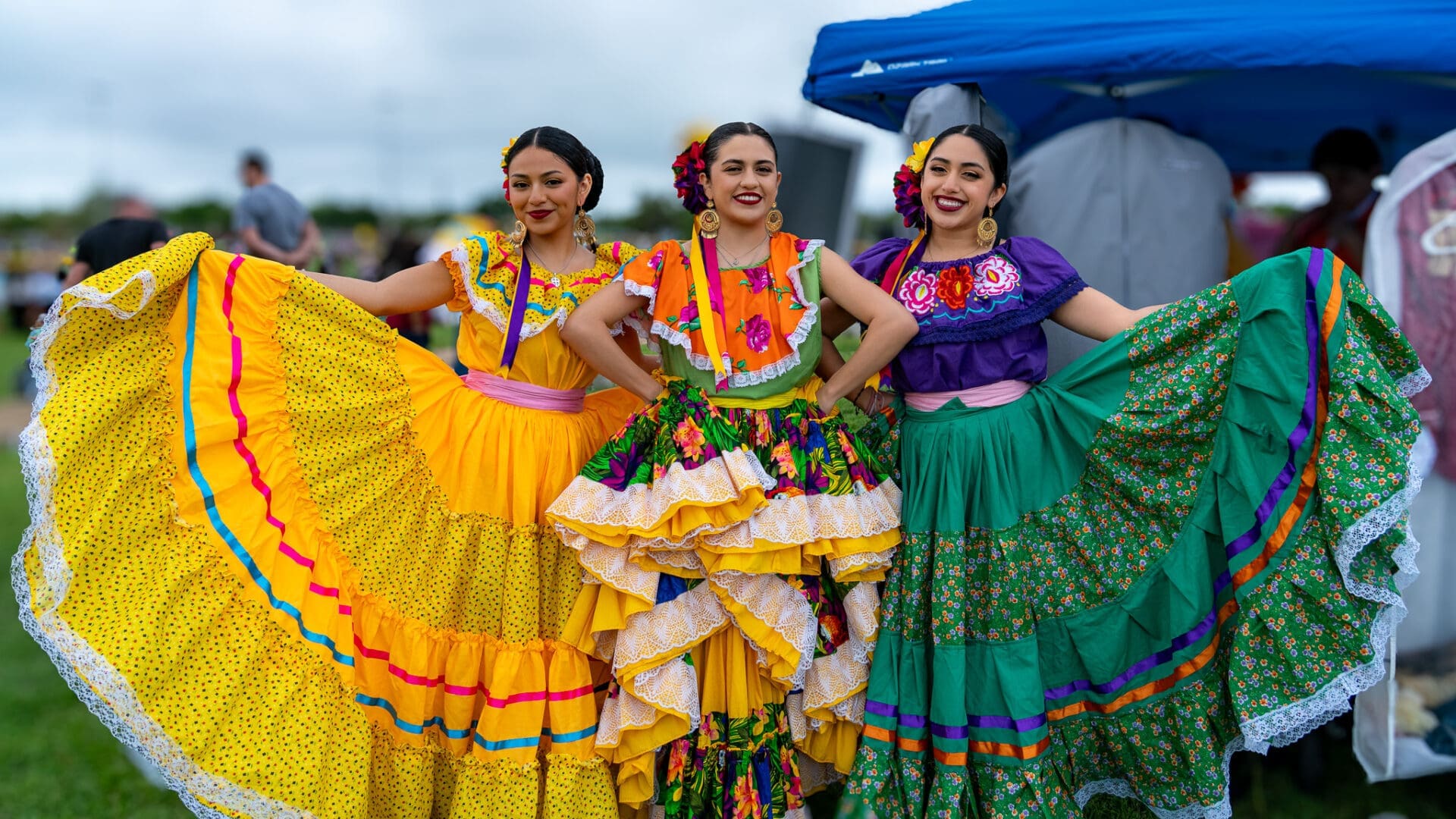 Three women stand side by side outdoors, smiling and wearing colorful traditional Mexican dresses with floral and ruffled patterns, holding out their skirts to display the vibrant fabric.
