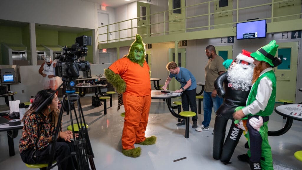 Multiple people stand in the middle of a cafeteria style room where a camera is pointed at the Grinch who is wearing an orange jumpsuit as though he is a member of the prison population. In the foreground another individual is dressed as a Leprechaun holding a wrestling dummy and a puppet while three other people stand with video equipment. 