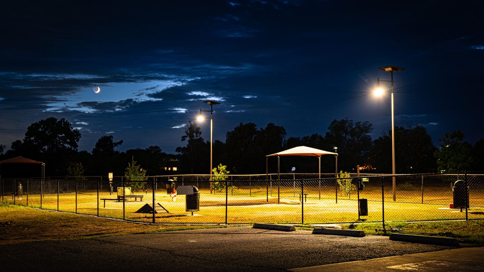 A fenced dog park at night, illuminated by tall lights, with a covered seating area and trees inside. The sky is dark blue with scattered clouds and a crescent moon visible.