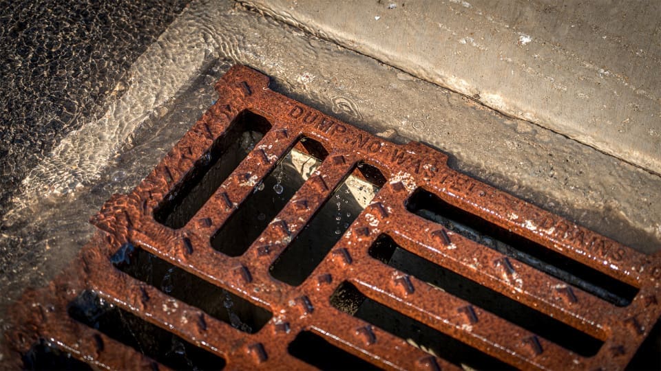 A close-up of water flowing into a rusty metal storm drain grate set in concrete. The grate is embossed with the words "DUMP NO WASTE.