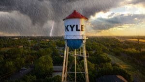 A rainstorm with lightning strikes near a tall water tower labeled "KYLE," standing above green trees and buildings, under a dramatic sky with dark clouds and sunlight breaking through.