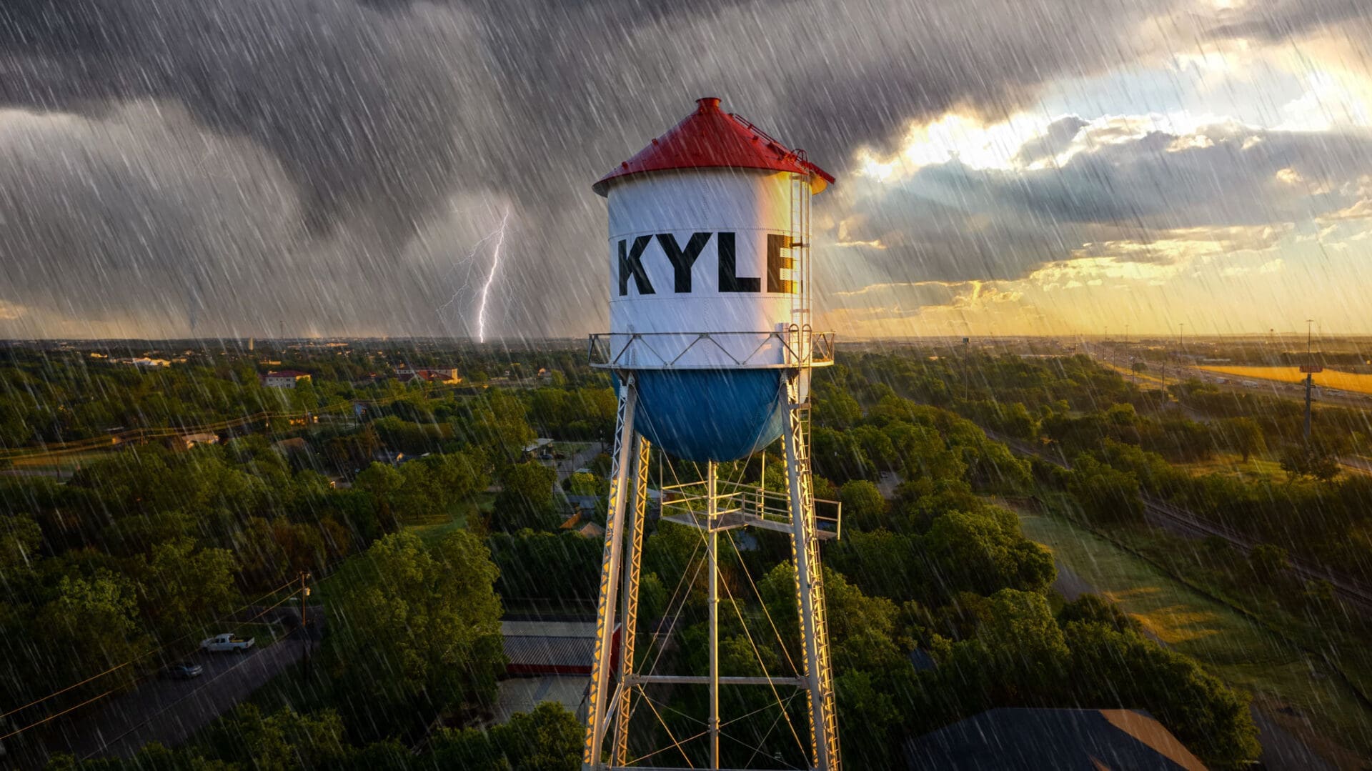 A rainstorm with lightning strikes near a tall water tower labeled "KYLE," standing above green trees and buildings, under a dramatic sky with dark clouds and sunlight breaking through.