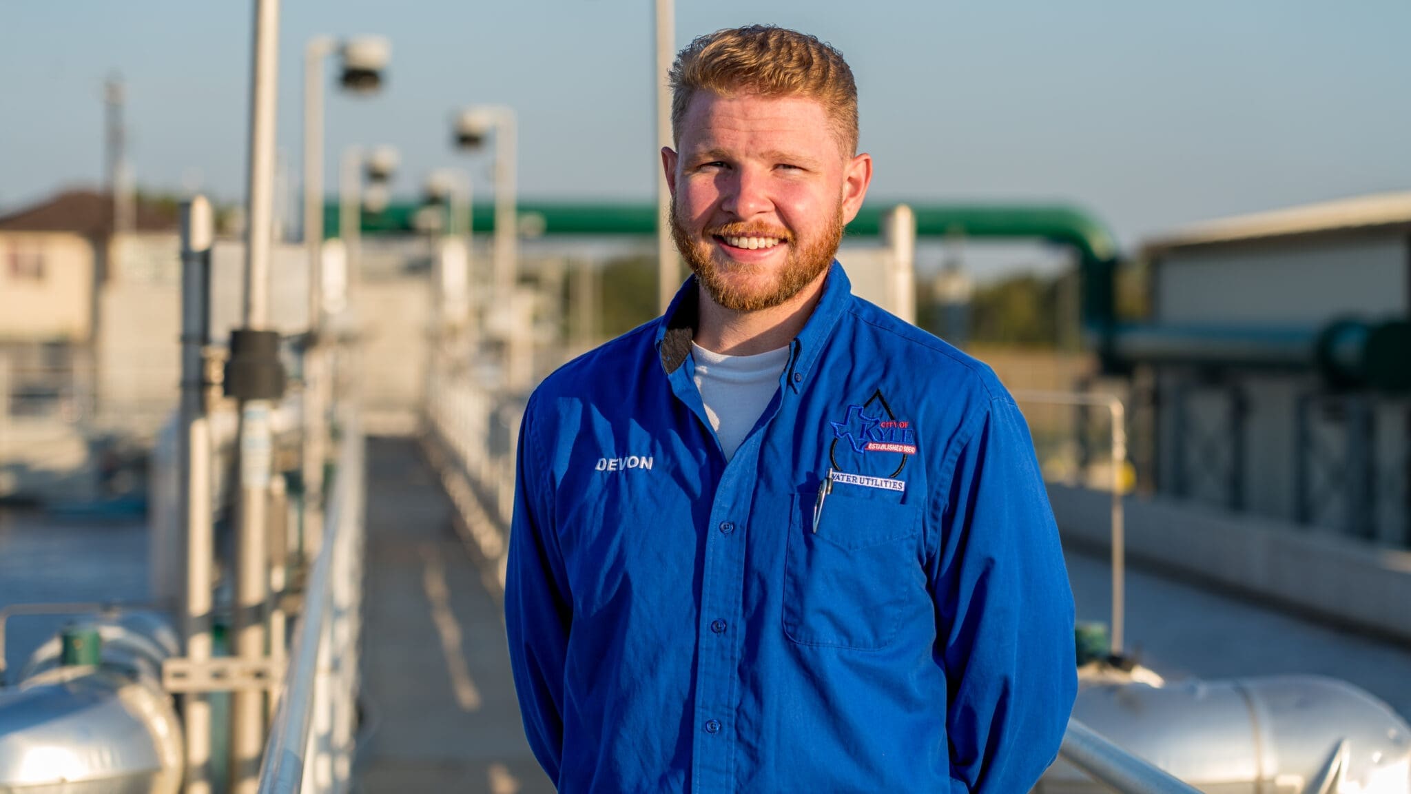 A man with short blond hair and a beard, wearing a blue work shirt, stands outdoors at a wastewater treatment facility, smiling at the camera. The background shows pipes and equipment in bright sunlight.