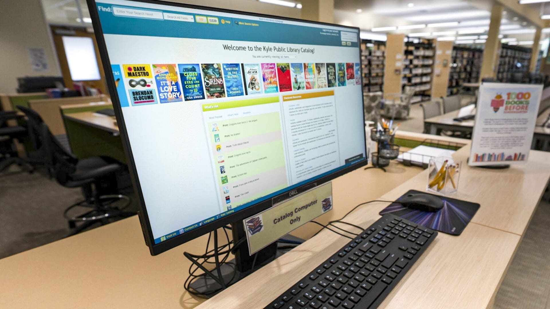 A computer displaying the library catalog screen is on a desk with a keyboard and mouse in a public library, surrounded by bookshelves and library signage.