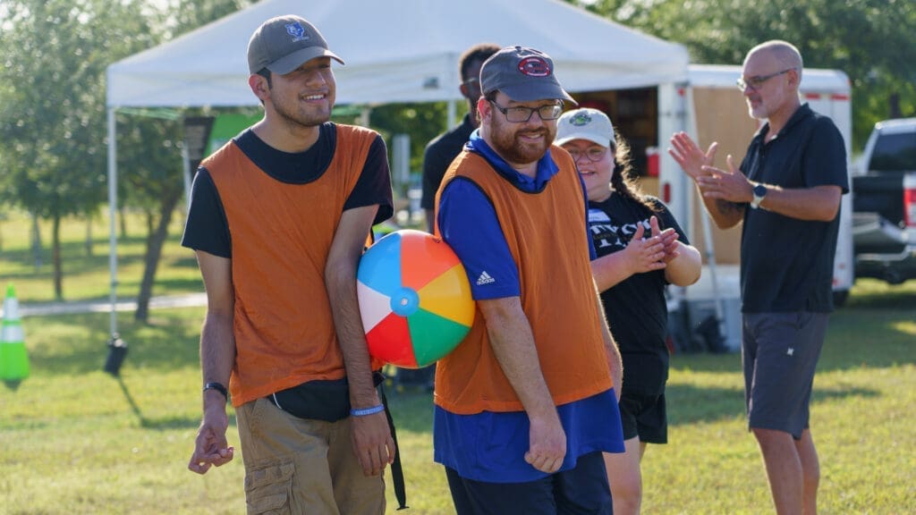 Two men wearing orange vests smile while holding a colorful beach ball between them at an outdoor event. Three people in the background clap and watch, with trees and a white tent visible.
